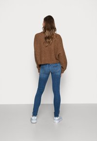 Suéter marrón de punto, corte holgado; jeans ajustados azules con un aspecto desvanecido; zapatillas de deporte azul claro que completan el conjunto, visto desde atrás.
