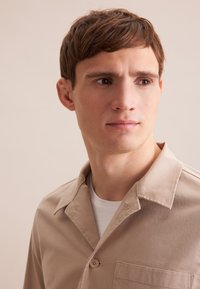 Camicia beige chiaro con design colletto, chiusura con un solo bottone e tasca applicata. Tessuto morbido con una texture liscia.