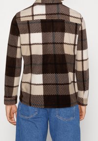 Camisa de forro polar de cuadros marrón y crema con cuello, mangas largas y una textura suave, que presenta diferentes patrones de cuadro y acentos.