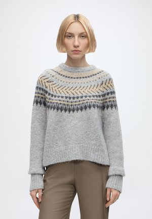 Jeune personne aux cheveux blonds courts portant un pull gris à motifs et un pantalon marron, debout devant un fond uni.