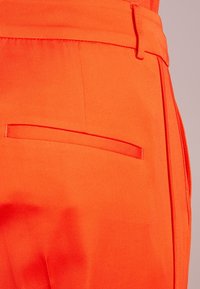 Jasnopomarańczowe spodnie z teksturowanej tkaniny, z dyskretną tylną kieszenią z poziomym szwem. Dobrze dopasowane, o gładkim wykończeniu.