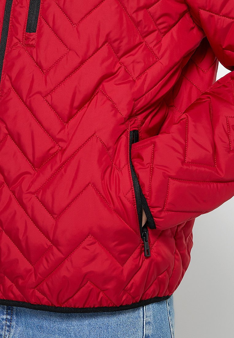 Giacca trapuntata rossa con motivo a zigzag, tasche con zip nere e orlo a costine. Materiale sintetico morbido con una texture gonfiata.