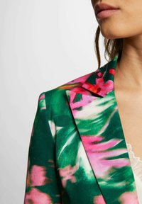 Blazer floreale caratterizzato da una combinazione di tonalità verdi e rosa, con una texture liscia e un colletto definito. Il design include ampi motivi di foglie astratte.