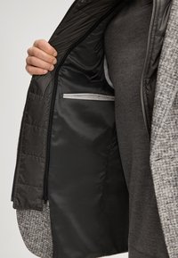 Grauer, strukturierter Außenmantel mit schwarzer, gesteppter Innenfütterung, ausgestattet mit einer inneren Reißverschlusstasche und glattem, reflektierendem Besatz. Hand greift die Öffnung.