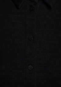 Zwarte overhemd met knopen, gemaakt van textuurstof, met een kraag en drie knopen aan de voorkant. Het materiaal heeft een subtiel gestreept patroon.