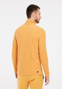 Pullover in pile di colore giallo senape, con colletto alto, maniche lunghe e una texture morbida, arricchito da un piccolo logo nero sul fondo.