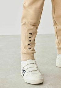 Beige sweatpants har et sort "BAKER" logo langs siden og en tekstureret panel på låret. Hvide sneakers med velcro-stropper og marineblå detaljer.