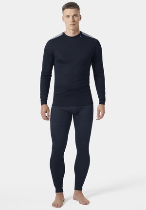 Mężczyzna stojący boso, ubrany w dopasowaną czarną koszulkę z długim rękawem oraz pasujące czarne legginsy na prostym białym tle.