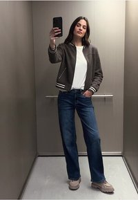 Šedá bomber bunda s bílými pruhovanými manžetami, bílá košile, modré džíny a béžové tenisky. Stojí v výtahu a drží telefon.
