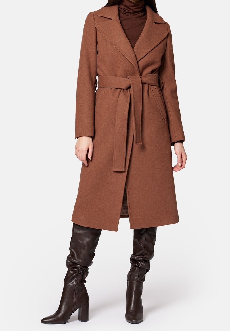 Femme portant un manteau marron ceinturé jusqu'aux genoux, par-dessus un col roulé marron foncé et des bottes marron foncé montantes à talons.