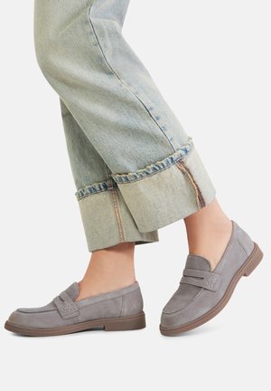 Jasnołówzane jeansy z podwiniętymi nogawkami w połączeniu z szarymi zamszowymi mokasynami wsuwanymi z brązową podeszwą na bosych stopach na jednolitym tle.