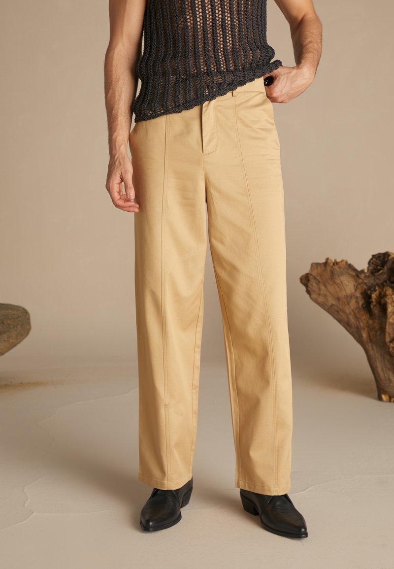 Homme portant un pantalon beige à jambes larges, des chaussures pointues noires et un haut sans manches en maille noire, debout sur un sol neutre.