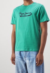 Camiseta de algodón verde con cuello redondo que presenta el logo de "Pepe Jeans" en azul oscuro y el texto "London Denim Brand" debajo. Corte casual.