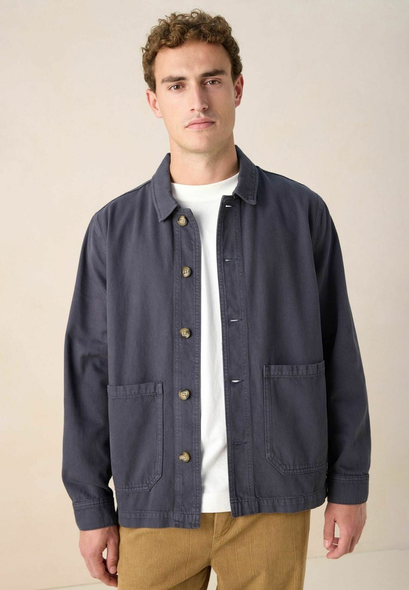 Donkerblauwe katoenen jas met een knoopsluiting aan de voorkant, twee voorzakken en een gekraagde halslijn. G gecombineerd met een wit shirt en corduroy broek.