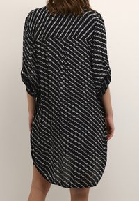 Femme portant une robe noire avec des motifs blancs diagonaux semblables à des chaînes, debout les bras détendus contre un fond clair uni.