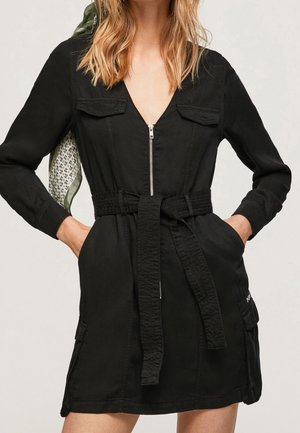 Mujer que lleva un vestido negro con cinturón, mangas largas, cremallera frontal y bolsillos con solapa, manos en los bolsillos laterales, con un pañuelo estampado en el hombro.