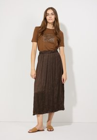 Bruine graphic t-shirt met korte mouwen, gecombineerd met een lange, geplooide bruine rok. Het model draagt tan, geweven slipper-sandalen. Eenvoudige achtergrond.