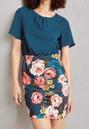 Frau trägt ein türkisfarbenes kurzärmliges Oberteil und einen hoch taillierten Rock mit rosa und gelben Blumenmustern.