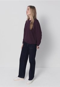 Dunkelrotes Langarmshirt kombiniert mit dunkelblauen Jeans mit geradem Bein und weißen Turnschuhen, vor einem hellen Hintergrund. Einfaches, lässiges Outfit.