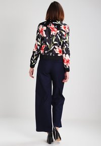 Veste bomber fleurie noire ornée de grandes lys roses ; poignets et ourlet côtelés. Associée à un pantalon large bleu marine et des chaussures noires.