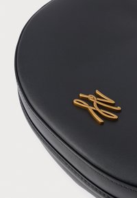 Kerek fekete bőr táska varrott szegéllyel, arany fonott fémbetűkkel a tetején, fehér háttér előtt.