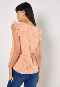 Top en cotton pêche avec un motif en œillets, des manches volantes et un ourlet festonné. Associé à un jean bleu, mettant en valeur une coupe décontractée et une texture.