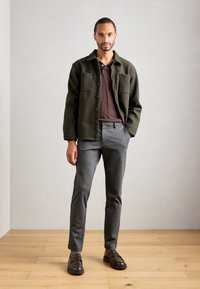 Hombre de pie en interiores, vistiendo una chaqueta verde oscuro, una camiseta polo marrón, pantalones grises y mocasines marrón oscuro sobre un suelo de madera.
