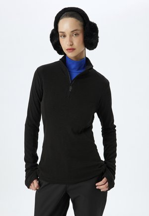 Zwarte lange mouwen top met een halflange rits kraag, gecombineerd met een blauwe high-neck onderlaag. Model draagt zwarte oorkappen en heeft zijn handen in de zakken.