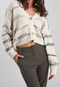 Cardigan lavorato a maglia color crema con strisce orizzontali grigie e beige, dotato di due bottoni di legno, indossato su pantaloni verde scuro.