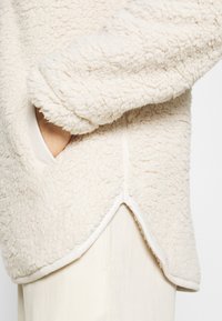 Käsi valkoisen, teksturoidun fleecejakun taskussa, jossa on halkiohelma, vaaleiden housujen päällä tasaisella taustalla.