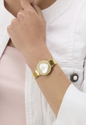 Ceas de mână aurit, cu cadran rotund alb, înconjurat de o ramă decorată cu cristale. Brățara are un design cu zale plate aurii.