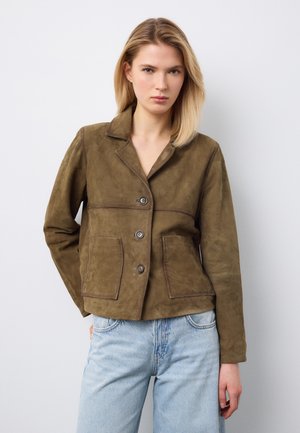 Mujer rubia con una chaqueta marrón de ante abotonada con dos bolsillos delanteros y vaqueros azul claro, posando contra un fondo blanco.