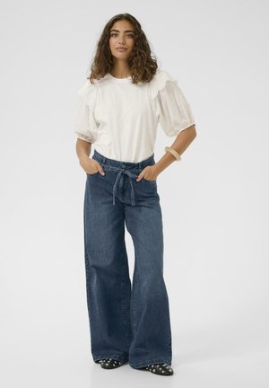 Moteris stovi su rankomis kišenėse, vilkėdama baltą palaidinę su pūstomis rankovėmis ir nėrinių detalėmis, platėjančias aukšto liemens džinsus ir juodus batelius su kniedėmis.