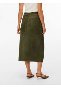 Femme portant une jupe midi vert olive avec des poches arrière, un haut blanc et des chaussures à talons bas marron à bride arrière, debout sur un fond blanc.