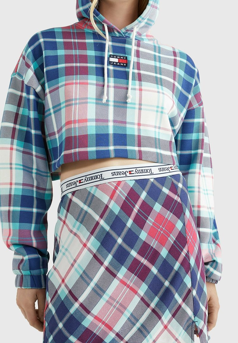 Felpa cropped con motivo a quadri multicolore, caratterizzata da tonalità di blu, rosa e bianco. Include una patch con logo e cappuccio con cordini. Gonna a quadri coordinata.