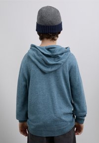 O hanorac albastru, texturat, cu glugă, mâneci lungi și tiv striat, asortat cu o căciulă tricotată gri, cu margine bleumarin.