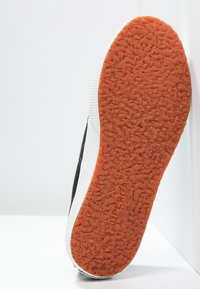 Suela de zapatilla en blanco y negro con una suela texturizada en naranja. Presenta un diseño plano y un patrón de agarre pronunciado. Marcada con "SUPERGA."