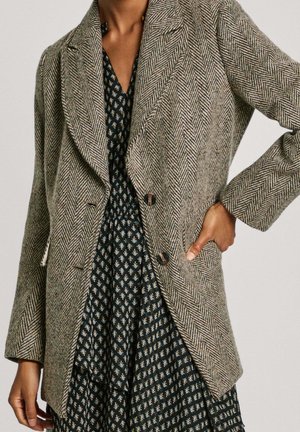 Frau trägt ein graues Fischgrät-Blazer mit braunen Knöpfen über einem schwarzen Kleid mit Blumenmuster, Hand in der Blazertasche.