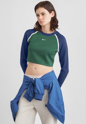 Frau, die einen grün-blauen, lässigen Nike-Sweatshirt und eine blaue Jacke um die Taille gebunden trägt sowie cremefarbene Hose.