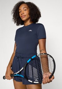 T-shirt atlético azul-marinho com mangas curtas, apresentando um pequeno logótipo branco. Acompanhado de calções azul-marinho, segurando uma raquete de ténis preta com detalhes azuis.