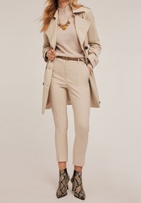 Trench-coat beige sur un pull léger, pantalon beige ajusté et bottines à imprimé serpent. Comprend une ceinture et un collier de perles colorées.