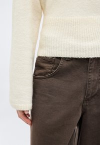 Άτομο που φοράει πλεκτό πουλόβερ κρεμ χρώματος και σκούρο καφέ παντελόνι, με το αριστερό χέρι να ακουμπά κοντά στην τσέπη του παντελονιού.