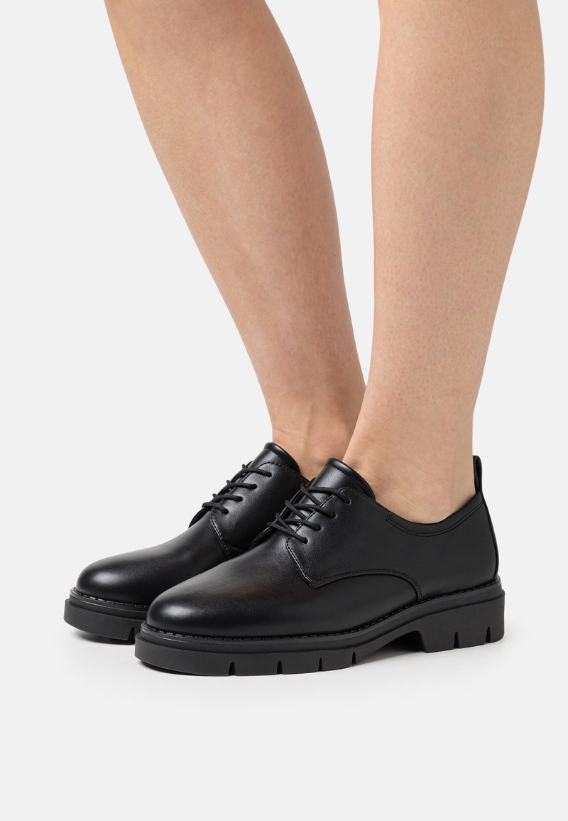 Jambes portant des chaussures en cuir noir à lacets avec des semelles en caoutchouc épaisses sur fond blanc, vues de profil et de face.