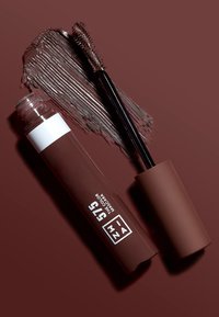 Tubo di mascara marrone con finitura opaca, dettagli bianchi, un applicatore e una striscia di prodotto marrone su uno sfondo simile.