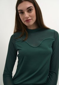 Dlouhý rukáv top v tmavě zelené látce s síťovým výstřihem ve vlnkovém vzoru, hladkou strukturou a vysokým límcem.