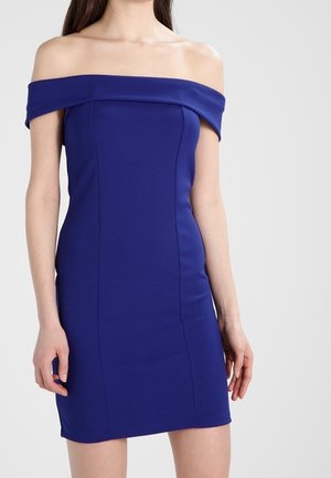 Vrouw draagt een nauwsluitende, koningsblauwe jurk met blote schouders en een korte zoom, staand tegen een effen lichte achtergrond.
