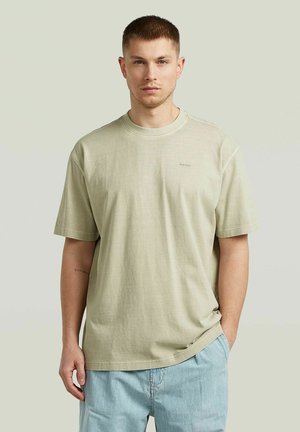 Young man with short brown hair wearing a light beige oversized t-shirt and light blue jeans standing with one hand in pocket.