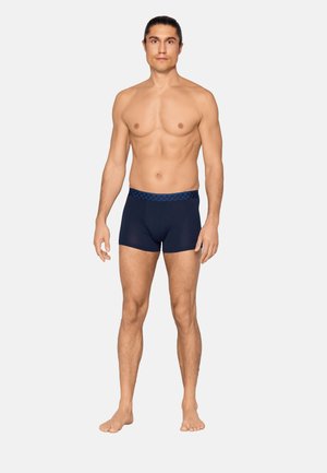 Hemdloser Mann steht barfuß und trägt marineblaue Boxer-Shorts mit gemustertem Bund vor schlichtem hellem Hintergrund.