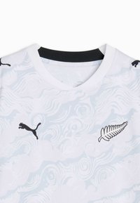 Maillot de sport blanc avec motifs abstraits tourbillonnants, logo Puma noir sur la poitrine gauche et emblème de fougère argentée sur la poitrine droite, doublure du col noire.