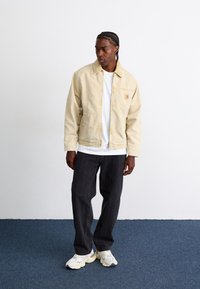 Veste zippée beige clair avec un col et une poche frontale, associée à un t-shirt blanc, un pantalon gris foncé et des baskets blanches.